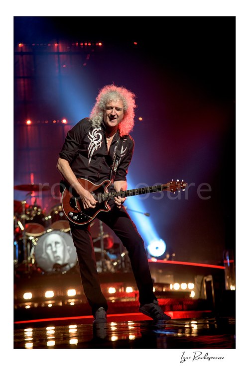 Vertical color image of Brian May of Queen playing guitar onstage under blue and red concert lighting with the drum kit visible behind him