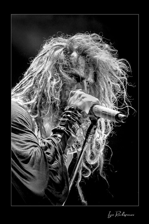 Vertical black and white image of Rob Zombie performing live on stage in a tight close-up with long dreadlocks, layered wristbands, and a handheld microphone