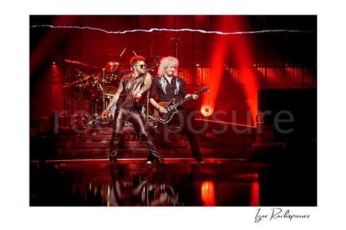 Horizontal color image of Adam Lambert and Brian May of Queen performing onstage together under vivid red concert lighting with the drummer visible behind them
