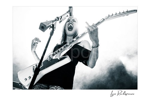 Horizontal black and white image of Lzzy Hale of Halestorm singing into a microphone while playing guitar in a dramatic low-angle live performance shot, with stage smoke rising behind her
