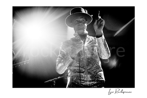 Gord Downie Tragically Hip Live Concert Print  Horizontal black and white image of Gord Downie of The Tragically Hip holding a microphone in a dramatic low-angle live performance shot, wearing a reflective jacket and hat beside an intense stage light burst
