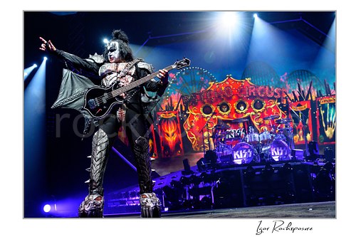 Gene Simmons of KISS performing in a wide power stance with bass guitar under bright white and blue stage lighting with fiery stage visuals behind him