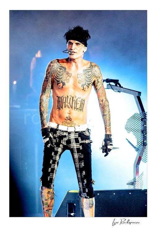 Vertical color image of Tommy Lee performing live during a Mötley Crüe concert, shirtless with tattoos and blue stage light