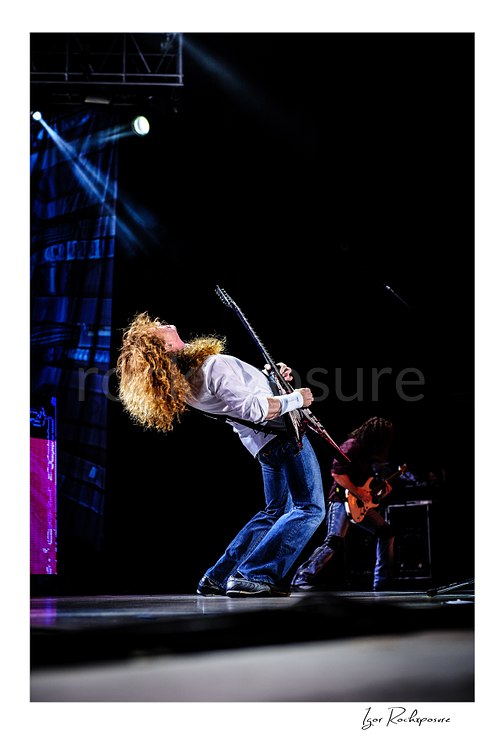 Vertical color image of Dave Mustaine of Megadeth leaning back while playing guitar on stage in a dramatic wide shot