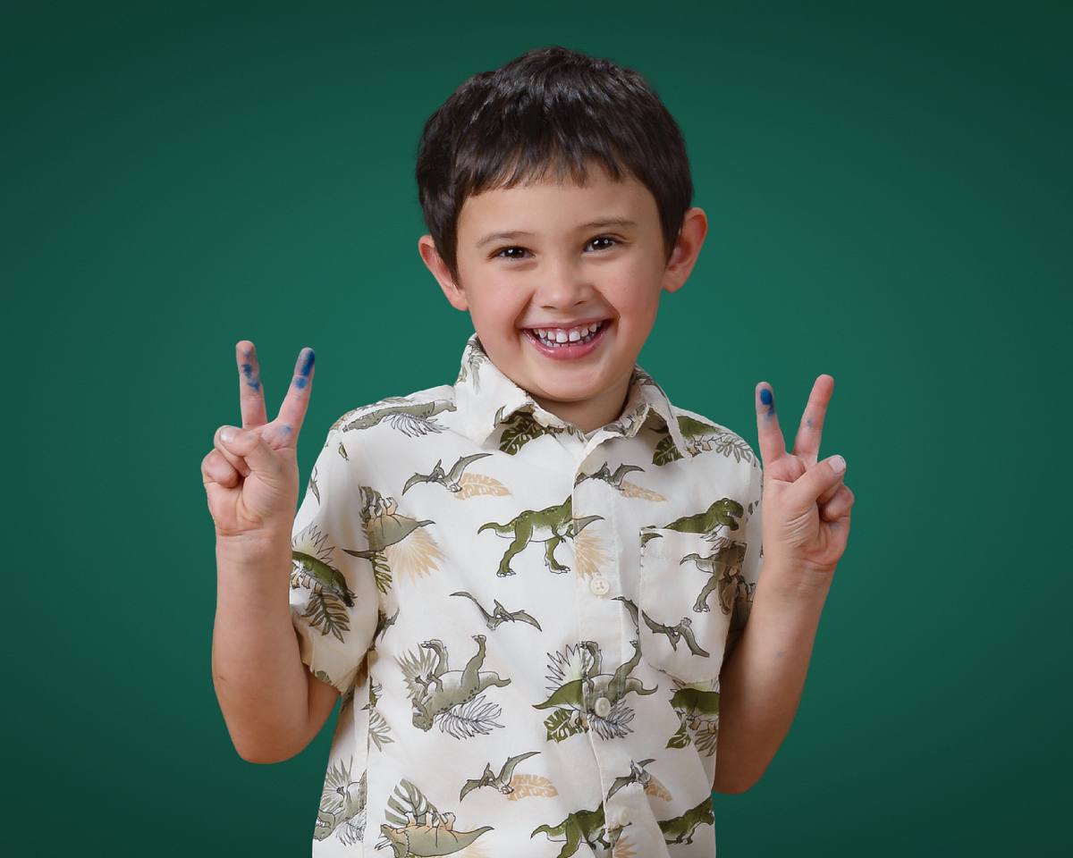 Preschool child making peace sign with painted fingers against green background