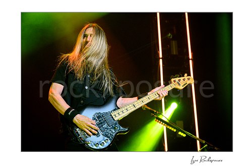 James LoMenzo of Megadeth plays bass with long blond hair across his face, green light behind him and warm vertical strips in the background.