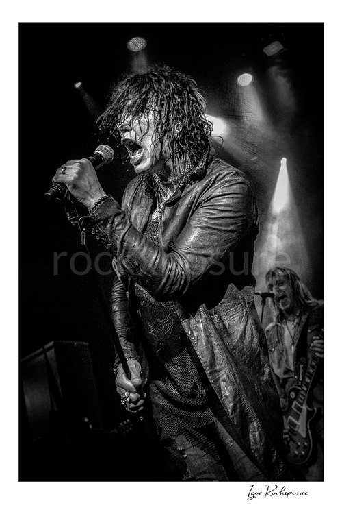 Vertical black and white image of Tom Keifer singing into a microphone in a fierce live performance close-up, with Jeff LaBar visible behind him under dramatic stage lights