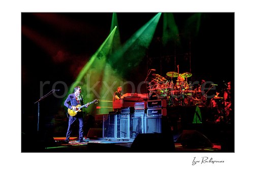Horizontal color image of Joe Bonamassa performing live on stage with his guitar under dramatic green and red lighting