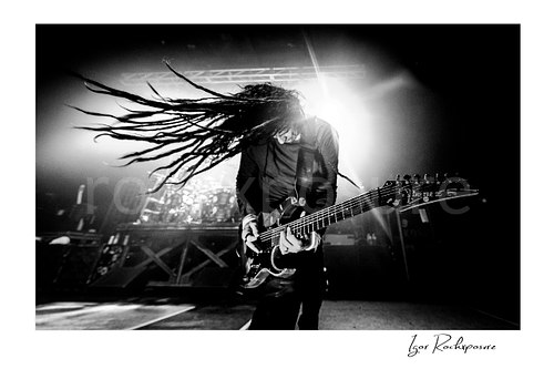 Horizontal black and white image of Korn guitarist James “Munky” Shaffer performing live on stage with long dreadlocks flying in dramatic motion