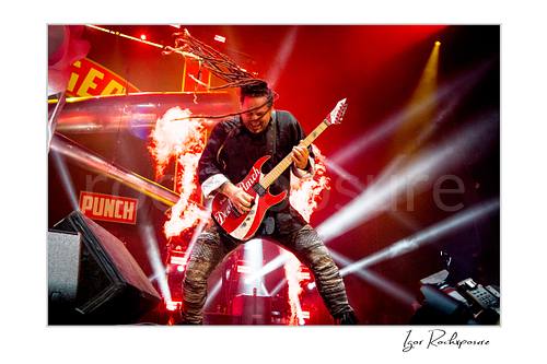 Horizontal color image of Jason Hook of Five Finger Death Punch playing guitar onstage with flying dreadlocks, flames, and bright concert beams