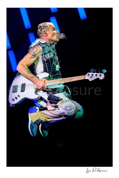 Flea hangs midair with both legs tucked behind his white bass, head turned sideways beneath glowing blue LED tubes against a black stage background.