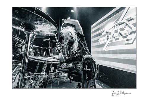 Horizontal black and white image of Robert Sweet of Stryper playing drums with raised drumsticks and flying hair