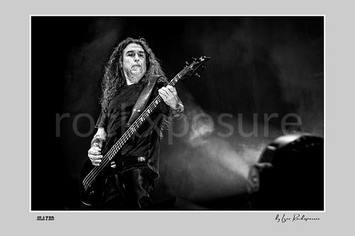 Horizontal black and white image of Tom Araya of Slayer playing bass onstage with long hair and drifting stage smoke