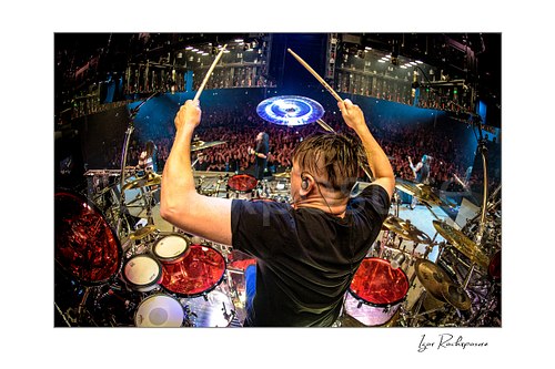 Horizontal color image of Korn drummer Ray Luzier seen from behind at the drums with drumsticks raised above the stage and crowd
