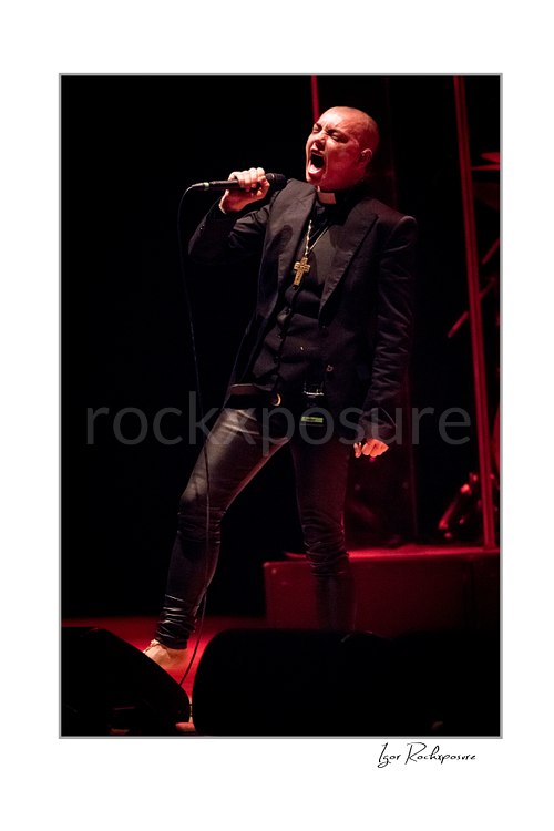 Sinéad O’Connor sings into a handheld microphone with a wide stance, wearing a black jacket and cross necklace against a dark stage edged with red light.