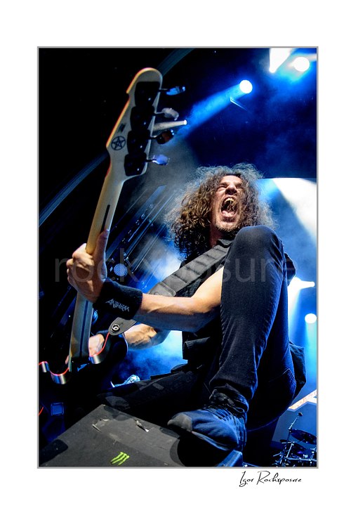 Vertical color low-angle image of Frank Bello of Anthrax performing live on stage, shouting while playing bass guitar under vivid blue concert lighting