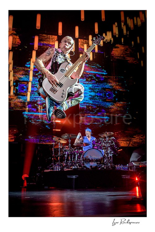 Flea jumps with both knees tucked beneath his white bass, high above Chad Smith’s drum kit against orange LED tubes and a colorful stage screen.