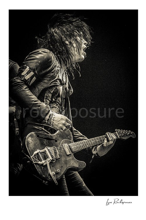 Vertical black and white image of Tom Keifer performing live on stage with an electric guitar, captured in side profile with wet hair and dramatic stage light against a dark background