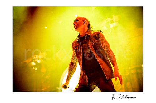 Horizontal color image of M. Shadows of Avenged Sevenfold on stage in a dramatic close-up with tattooed arms, sunglasses, and vivid yellow light