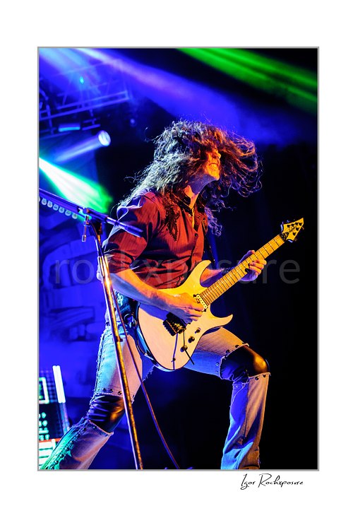 Vertical color image of Kiko Loureiro of Megadeth playing guitar on stage with flying hair and vivid blue and green stage lights