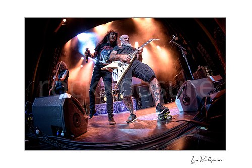 Horizontal color image of Joey Belladonna and Scott Ian of Anthrax performing live on stage, with Belladonna at left holding a microphone and Scott Ian at right playing guitar under warm concert lighting
