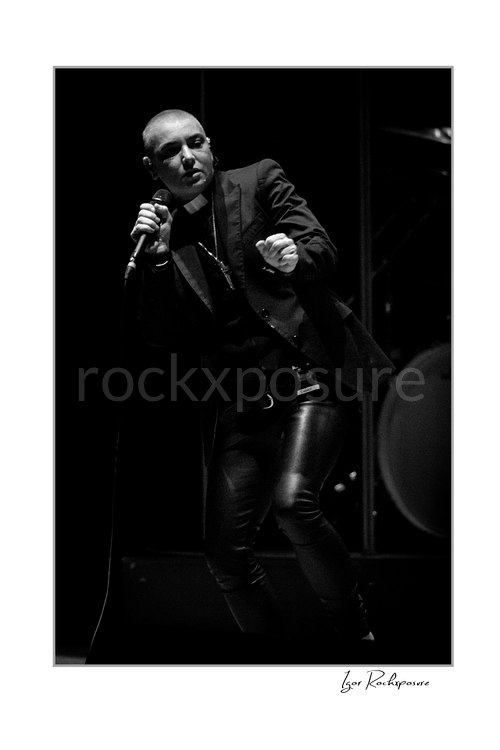 Sinéad O’Connor leans sideways with bent knees, holding a microphone near her face and her free hand forward in a black and white stage portrait.