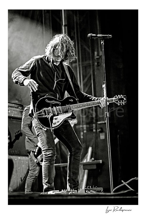Vertical black and white image of Chris Cornell performing live on stage, playing electric guitar beside a microphone stand under concert lighting
