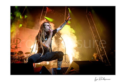 Horizontal color image of Randy Blythe of Lamb of God crouched on stage with long dreadlocks, handheld microphone, and a burst of fire behind him