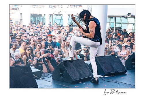 Horizontal color image of Nuno Bettencourt performing live on stage with an electric guitar, one foot raised on a stage monitor before a large crowd