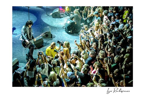 Horizontal color image of Tom Keifer performing live at the edge of the stage with a black guitar, viewed from above as a packed crowd reaches toward him with raised hands and cameras