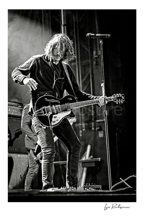 Vertical black and white image of Chris Cornell performing live on stage, playing electric guitar beside a microphone stand under concert lighting