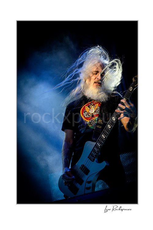 Vertical color image of John Campbell of Lamb of God playing bass on stage with flying white hair and blue stage smoke
