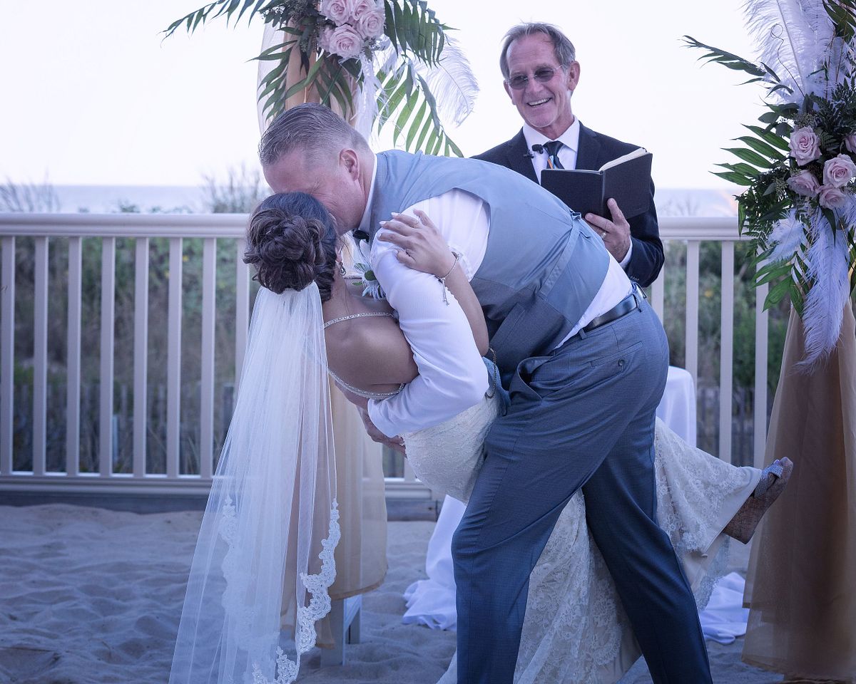 groom dipping his new wife under the wedding arbor after the ceremony on the beach in sussex county de, the minister is smiling