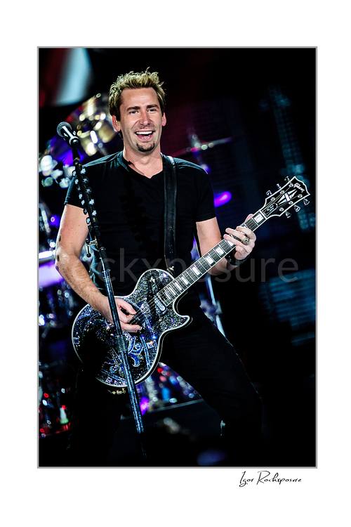 Vertical color image of Chad Kroeger of Nickelback playing guitar onstage beside a microphone stand with the drummer visible behind him