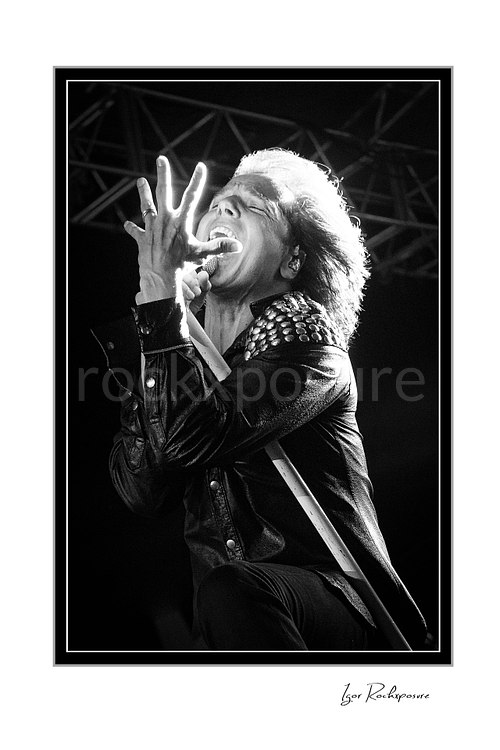 Vertical black and white image of Joey Tempest of Europe performing live on stage with a white microphone stand, studded black jacket, bright backlight, and expressive singing pose