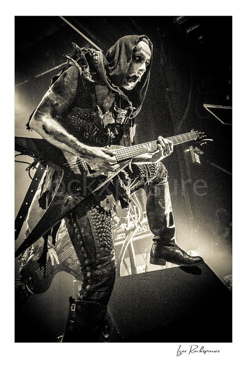 Vertical black and white image of Nergal of Behemoth playing guitar with one boot raised in a dramatic low-angle live performance shot