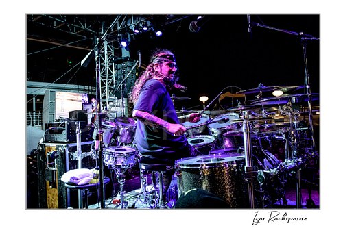Horizontal color image of Mike Portnoy playing drums from the side under purple stage lighting on a nighttime outdoor stage