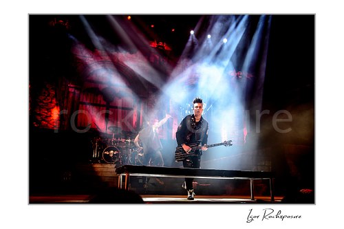 Horizontal color image of Johnny Christ of Avenged Sevenfold playing bass on stage beneath dramatic white spotlights and stage smoke