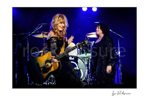 Horizontal color image of Nancy Wilson and Ann Wilson performing live on stage during a Heart concert with acoustic guitar and blue stage light