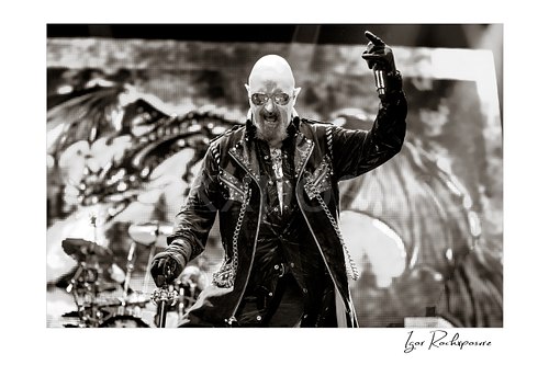 Horizontal black and white image of Rob Halford of Judas Priest performing live on stage, holding a microphone and cane in front of a dramatic LED screen backdrop