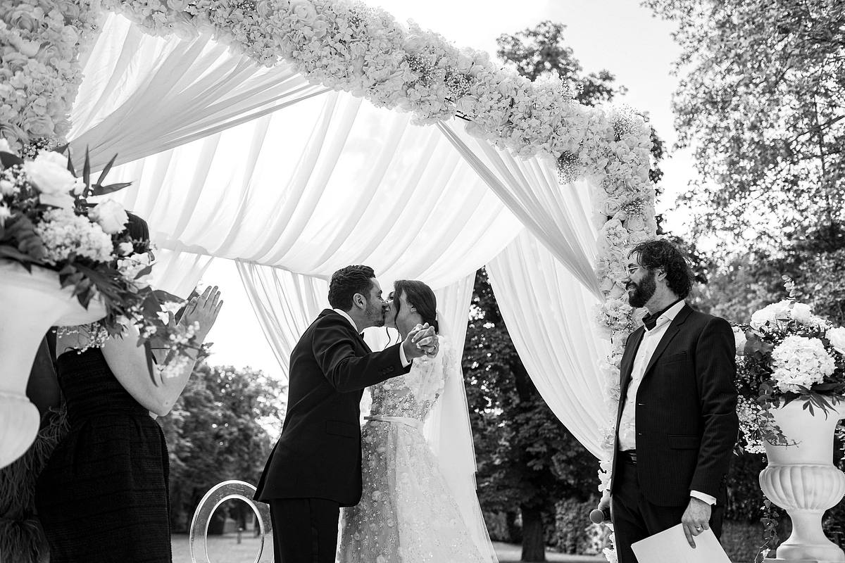Sebastien CLAVEL Photographe mariage Lyon Le couple de jeunes mari&eacute;s marche main dans la main sur un tapis blanc lors de leur c&eacute;r&eacute;monie de mariage en plein air, entour&eacute; d'invit&eacute;s et de d&eacute;corations florales