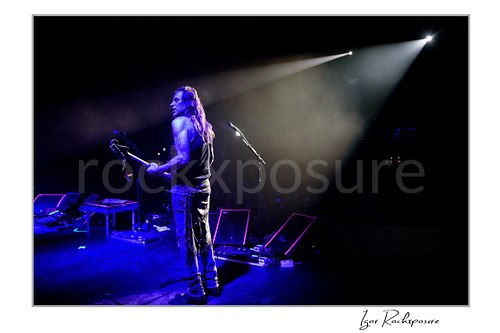 Horizontal color image of Nuno Bettencourt standing on stage with a guitar under dramatic blue light and a bright spotlight