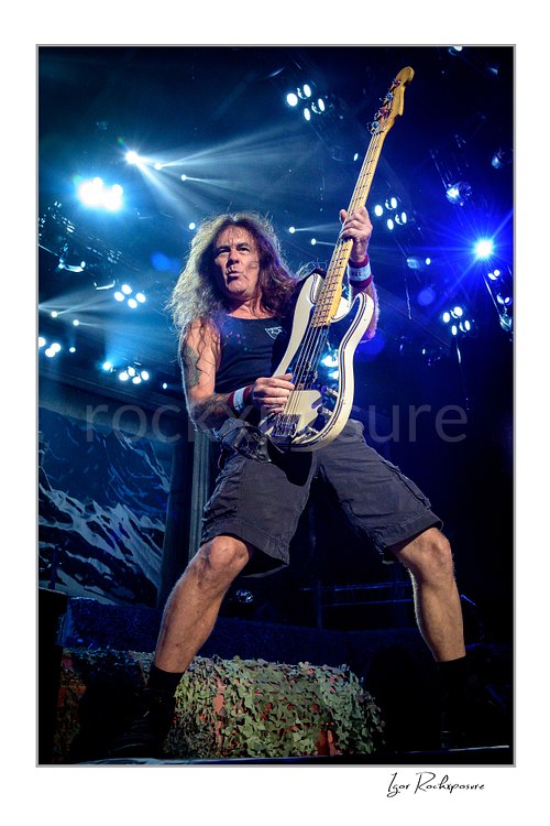 Vertical color image of Steve Harris of Iron Maiden performing live on stage, playing bass guitar in a wide stance under bright blue concert lights