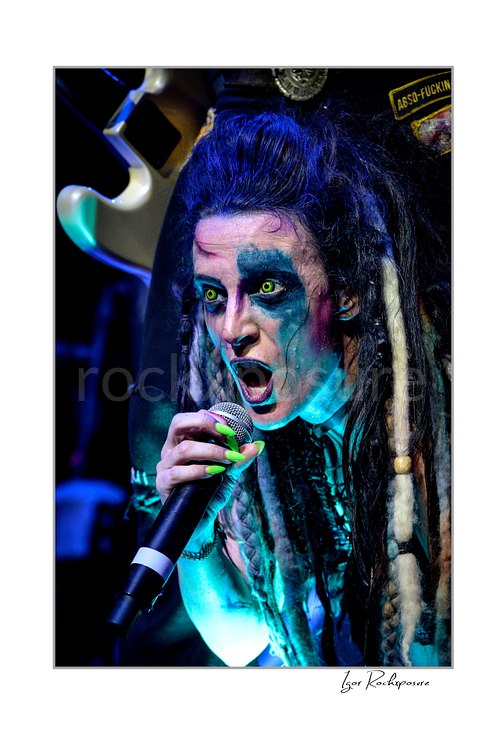 Vertical color image of Calico Cooper of Beasto Blanco singing into a microphone in an intense live performance close-up, with dramatic eye makeup, dreadlocks, and neon green nails under blue stage light