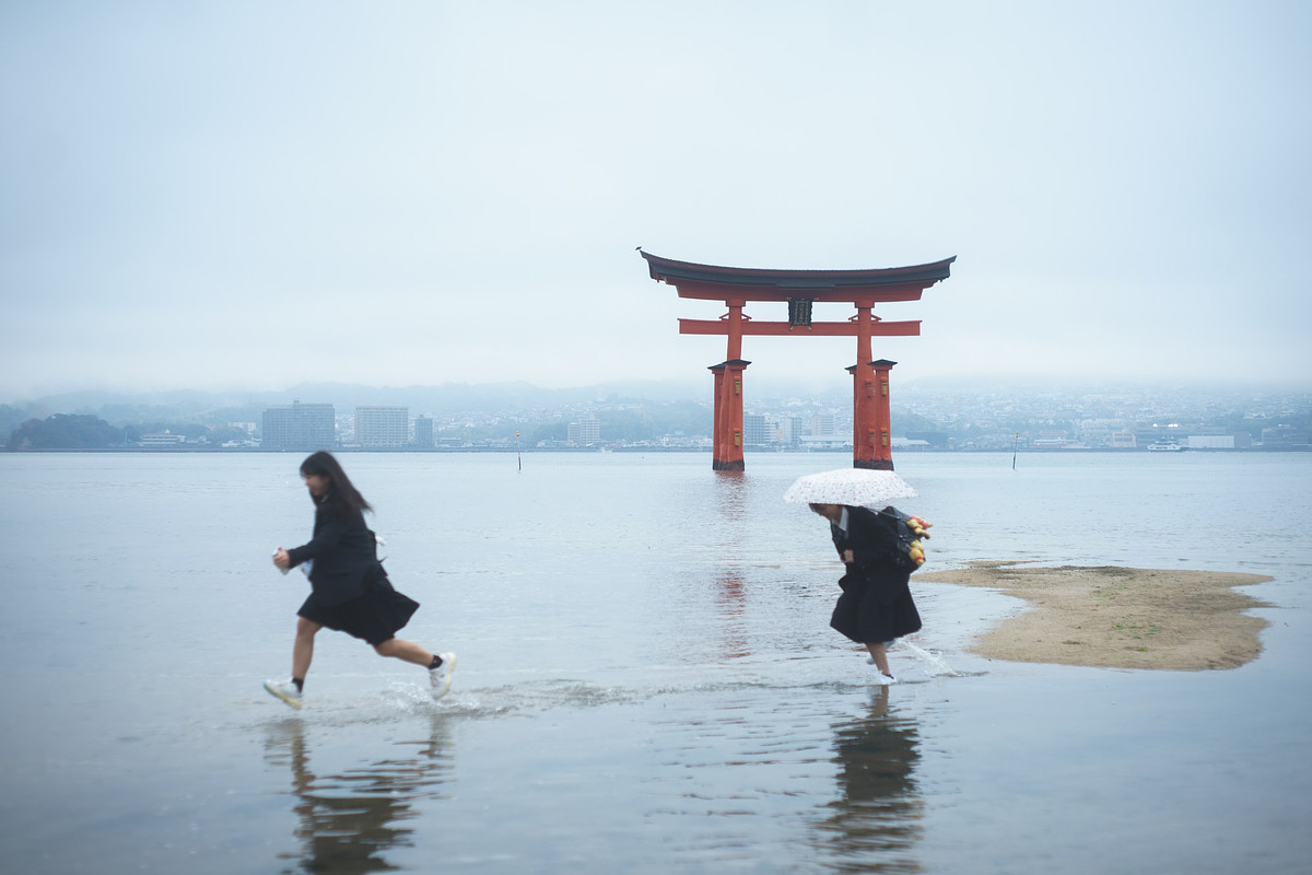 Torii & Sea : deux écolières traversent l'eau en courant devant le torii d'Itsukushima à marée montante, Miyajima