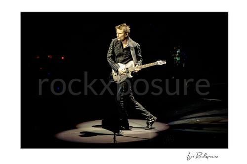 Horizontal color image of Matt Bellamy of Muse playing guitar alone in a spotlight on a dark stage
