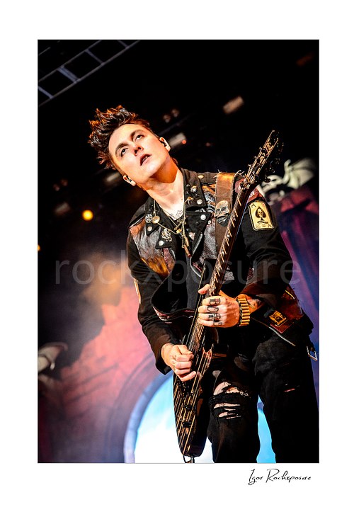 Vertical color image of Synyster Gates of Avenged Sevenfold playing guitar onstage under warm concert light and light stage haze