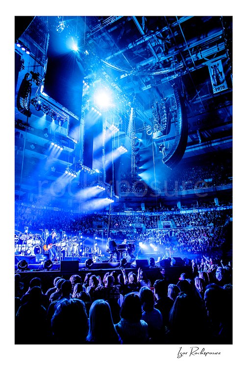 Vertical color image of Billy Joel performing on stage in a dramatic wide arena view with bright blue lighting and a large crowd