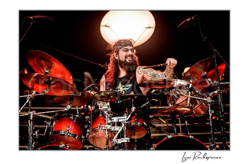 Horizontal color image of Mike Portnoy smiling at the drum kit with a drumstick raised under warm red stage lighting and a glowing round light behind him
