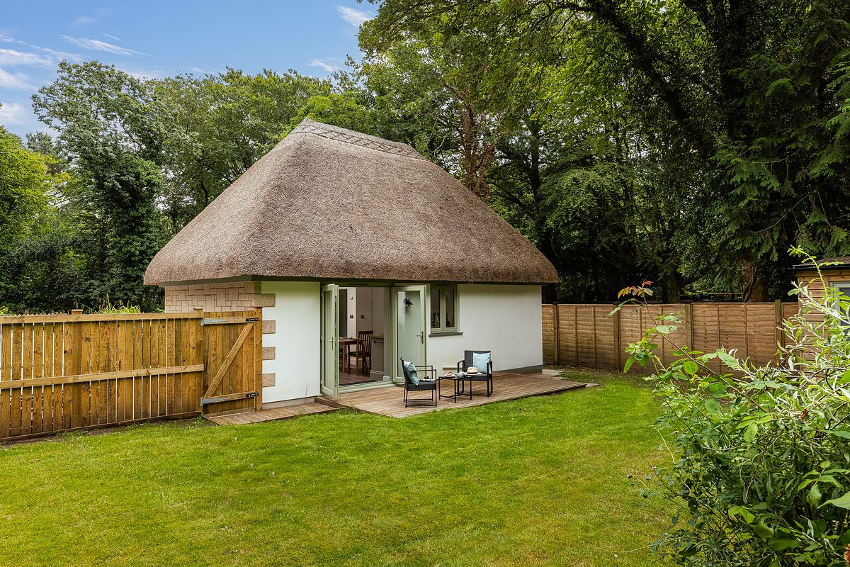 Small white cottage with a thatched roof, patio seating area, and fenced yard surrounded by lush green trees and grass.